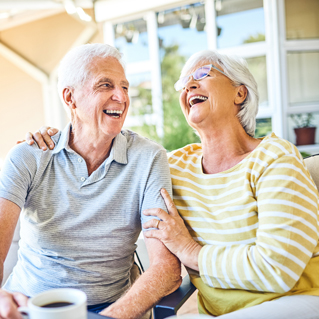 An older man and woman sit together, smiling and laughing. The man holds a coffee mug, while the woman rests her hand on his shoulder. They appear happy and relaxed, enjoying each other's company on a bright day.