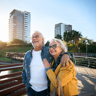 An elderly couple smiles and embraces while standing on a bridge, with modern buildings, trees, and a bright blue sky in the background.
