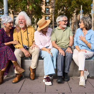 Five older adults sit together on a bench outdoors, smiling and laughing. They appear relaxed and happy, enjoying each other's company on a sunny day, with trees and cityscape in the background.
