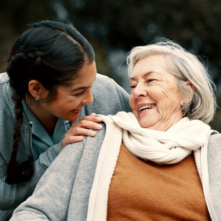 A young woman smiles warmly and places her hand on the shoulder of an elderly woman, who is laughing. They are outdoors, dressed warmly, and share a moment of joy and connection.
