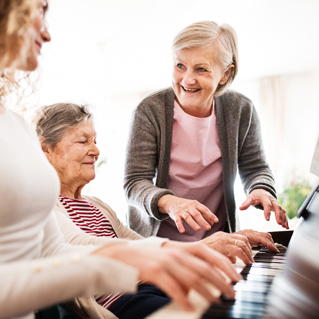 Three women, two elderly and one younger, smiling and playing piano together indoors. One woman stands, appearing to teach or guide, while the other two sit and play. The scene is bright and cheerful.