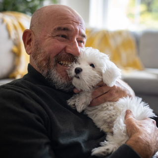 A smiling older man with a beard cuddles a small, fluffy white dog while sitting on a couch indoors. The background is softly lit with a yellow-patterned blanket visible.