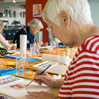 An elderly woman with short white hair and glasses, wearing a red and white striped shirt, paints a flower at a table in an art class with other seniors in the background. Art supplies and water glasses are on the table.