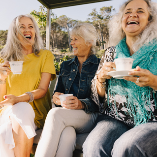 Three older women with gray hair are sitting outdoors, laughing and holding teacups. They are dressed casually and appear to be enjoying a cheerful conversation on a sunny day.