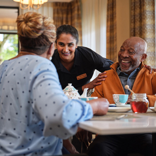 A smiling caregiver stands beside an elderly man and woman who are sitting at a table, enjoying tea together in a bright, cozy room. Everyone appears happy and engaged in friendly conversation.