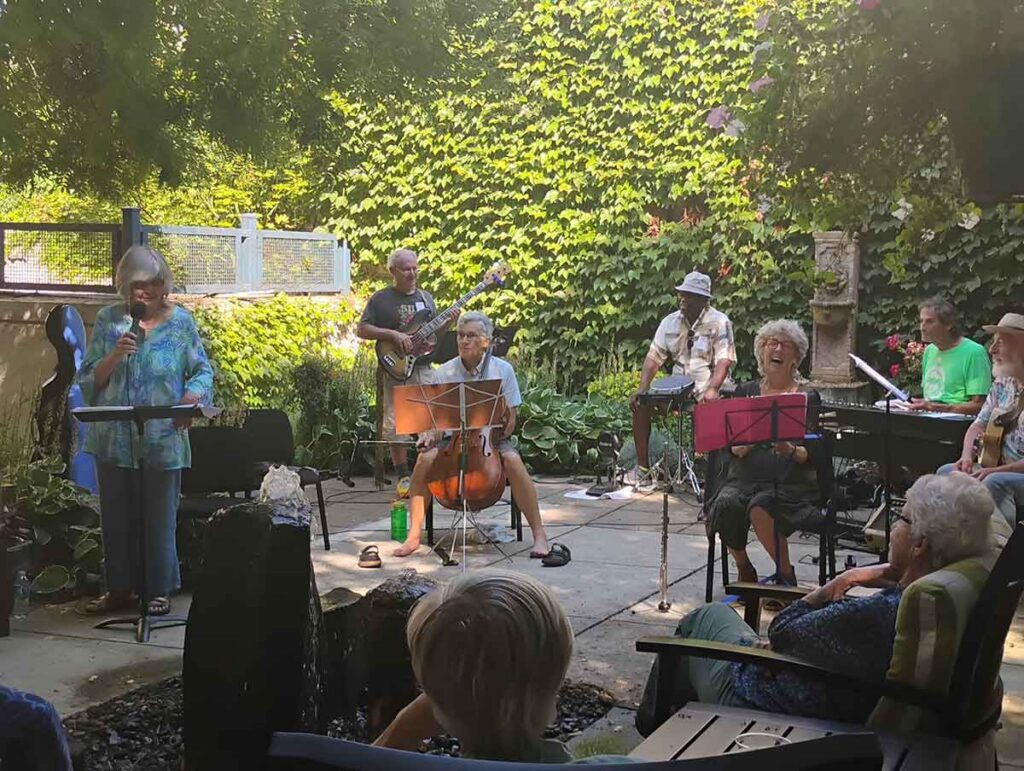 A small band performs outdoors in a leafy garden. Musicians play guitar, cello, keyboard, flute, and other instruments, while an audience watches from chairs. Sunlight filters through the trees.