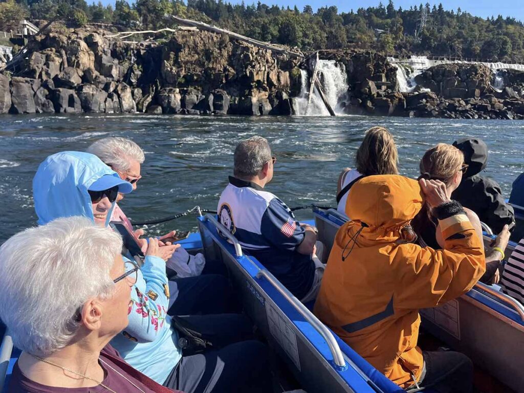 A group of people in a boat look toward a rocky waterfall and lush green trees under a clear sky. Some wear jackets and sunglasses, enjoying the scenic view on the water.