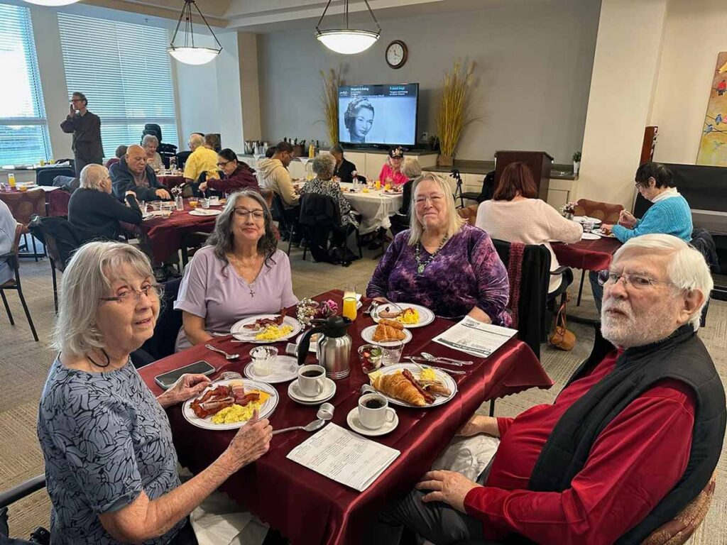 A group of older adults sit at a table enjoying breakfast in a busy dining room. Plates of food and coffee cups are on the table. Other people are dining and a TV screen is visible in the background.