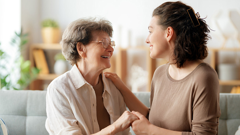 An elderly woman and a young woman sit together on a couch, smiling and holding hands warmly, sharing a happy moment in a bright, cozy living room—an ideal setting for learning how to talk to aging parents about their future.