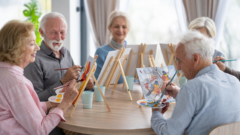 A group of older adults sit around a table, smiling and painting on canvases set on easels. The cheerful, social atmosphere offers a comfortable space—perfect for learning how to talk to aging parents about their future.