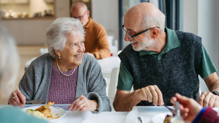 Two elderly people sit together at a table, smiling and talking while enjoying a meal as part of senior living activities. Other people are visible in the background, also dining in a bright, communal setting.